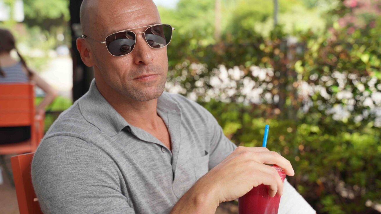 man holding a smoothie sitting at table with bushes in background