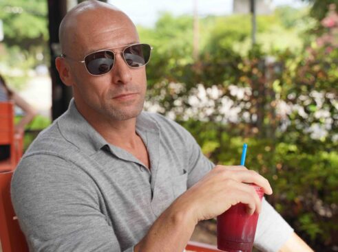 man holding a smoothie sitting at table with bushes in background